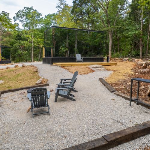 Outdoor seating area with Adirondack chairs on a gravel path, surrounded by trees, with a small table and a wooden platform nearby.