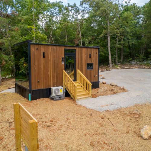 A small, modern cabin in the woods with wooden siding and a porch, surrounded by trees, and a concrete driveway leading to it.