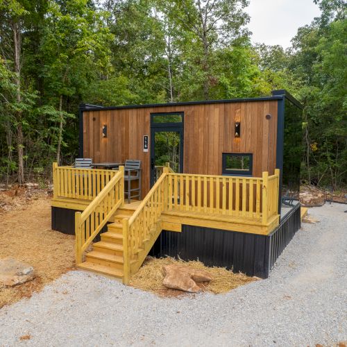 A small wooden cabin with a deck and stairs is surrounded by trees and a gravel path, nestled in a forested area.