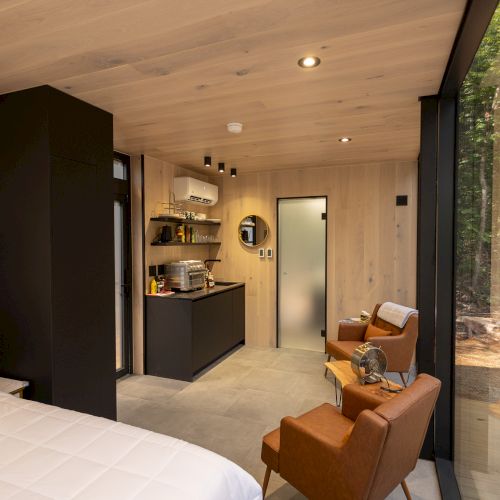 A cozy cabin interior with a bed, kitchenette, two chairs, and large window overlooking a forested area.