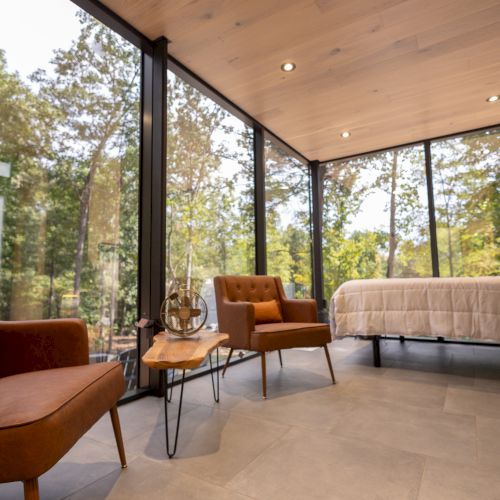 The image shows a modern room with glass walls, two brown chairs, a small table, and a bed, offering a view of trees outside.