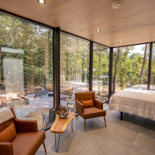 A modern room with glass walls features two chairs, a small table, and a bed, offering a scenic view of the surrounding forest.