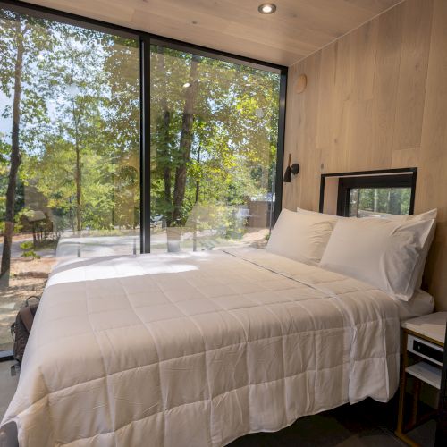A cozy bedroom with a white bedspread, large glass windows, and a view of a forest. The room has a minimalistic, tranquil design.