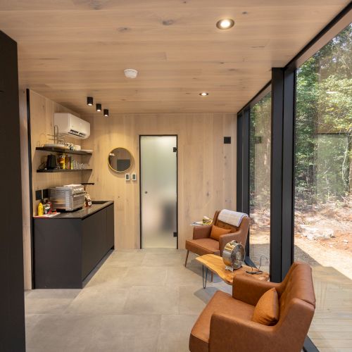 A cozy cabin interior with modern kitchen, leather chairs, large windows showing a forest view, and wooden walls; inviting and minimalist.