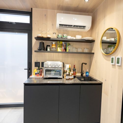 A compact kitchen with a countertop, toaster oven, shelves with kitchenware, a sink, air conditioning unit, and a round mirror on the wall.