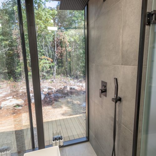 A modern bathroom with a glass shower, sleek sink, and a large window providing a view of the forest outside.