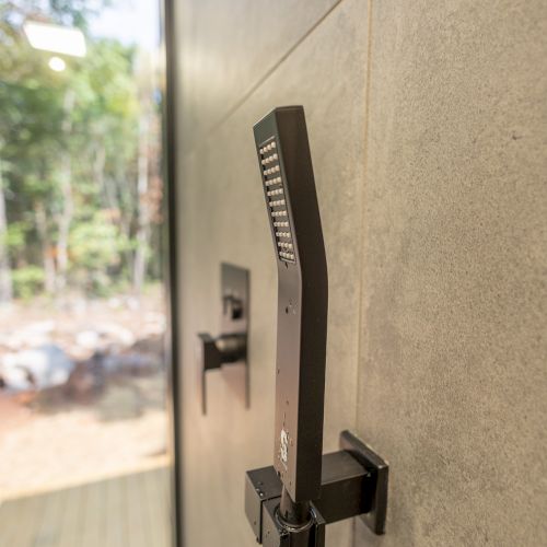 The image shows a modern showerhead mounted on a tiled wall, with a view of trees through a window in the background.