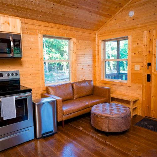 Cozy wooden cabin interior with a kitchenette, leather couch, and round ottoman, featuring a stove, microwave, and large windows.