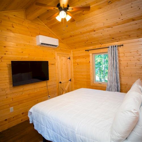 A cozy wooden bedroom with a bed, wall-mounted TV, air conditioning, window, white bedding, and ceiling fan, creating a warm, inviting atmosphere.