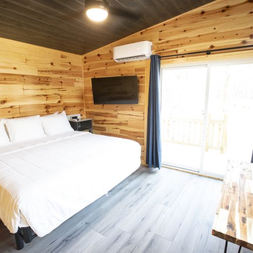 The image shows a cozy room with a wooden interior, a large bed, wall-mounted TV, and a sliding glass door leading outside.