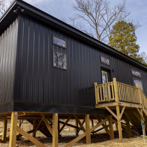 A small black elevated cabin with wooden stairs and windows, surrounded by trees under a blue sky.