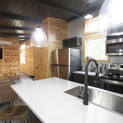 A modern kitchen with a white island, black faucet, stainless steel appliances, and wooden accents. Cozy living area visible in the back.