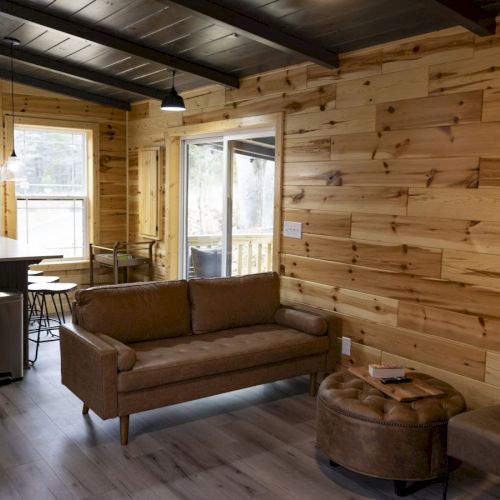 The image shows a cozy room with wooden walls, featuring a sofa, round ottoman, and a small dining area near a window in the background.