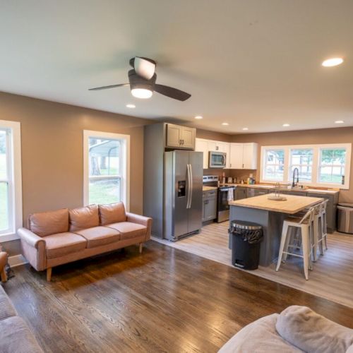 A modern open-concept living room and kitchen with a beige couch, island with stools, stainless steel fridge, and hardwood floors.