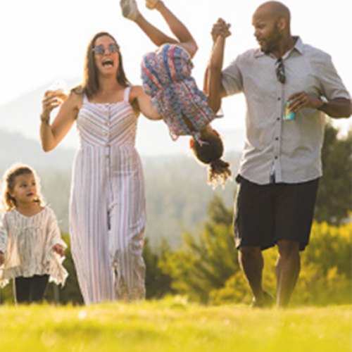 A family of four enjoys the outdoors, with the parents playfully swinging one child in the air while the other child walks alongside them.