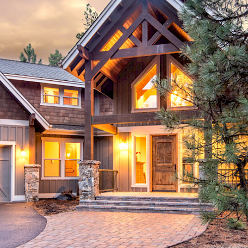 A large, two-story house with warm exterior lighting, multiple windows, a wooden facade, and a spacious driveway, surrounded by trees and a forest.