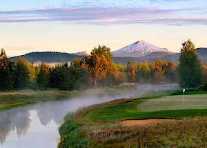 A serene landscape featuring a golf course, a river with mist, lush trees, and a snow-capped mountain in the background under a clear sky.
