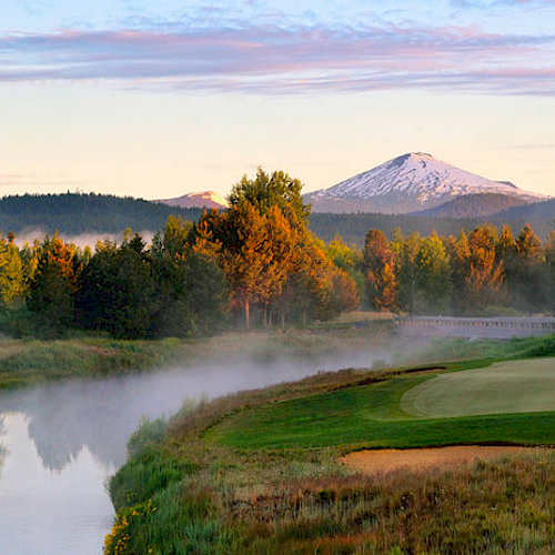 A serene landscape featuring a golf course, a river with mist, lush trees, and a snow-capped mountain in the background under a clear sky.
