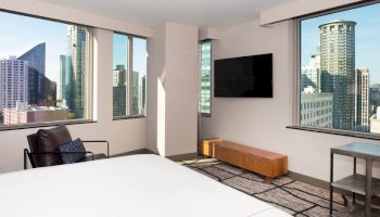 A modern hotel room with large windows offering a cityscape view, a bed, a chair, a TV mounted on the wall, and simple furniture ending the sentence.