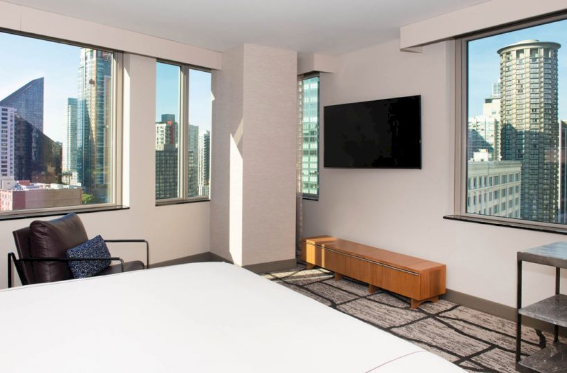 A modern hotel room with large windows offering a cityscape view, a bed, a chair, a TV mounted on the wall, and simple furniture ending the sentence.