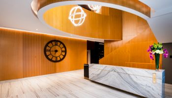 A modern lobby with a marble reception desk, wooden panels, large clock, spiral staircase, and hanging lights, plus a vase of flowers.