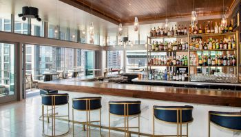 A modern bar with high stools and a wide array of liquor bottles on display, with large windows overlooking the city.