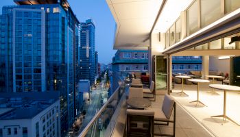 A modern cityscape view from a balcony at dusk. The balcony features outdoor seating, tables, and is adjacent to an indoor lounge area with large windows.