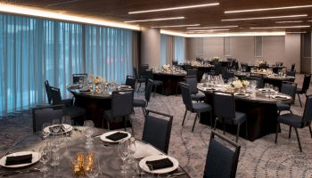 A dining room with round tables set for a formal event, featuring black chairs, neatly arranged table settings, and dim ambient lighting.