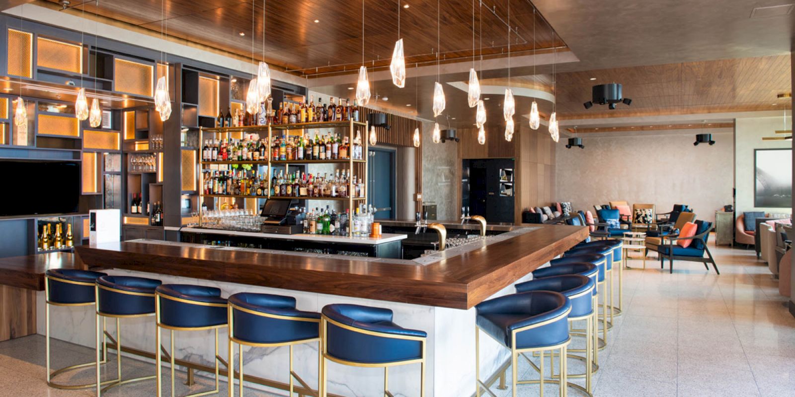 The image shows a modern bar with wooden accents, blue barstools, and hanging lights. The bar is stocked with various bottles, and there's a seating area.