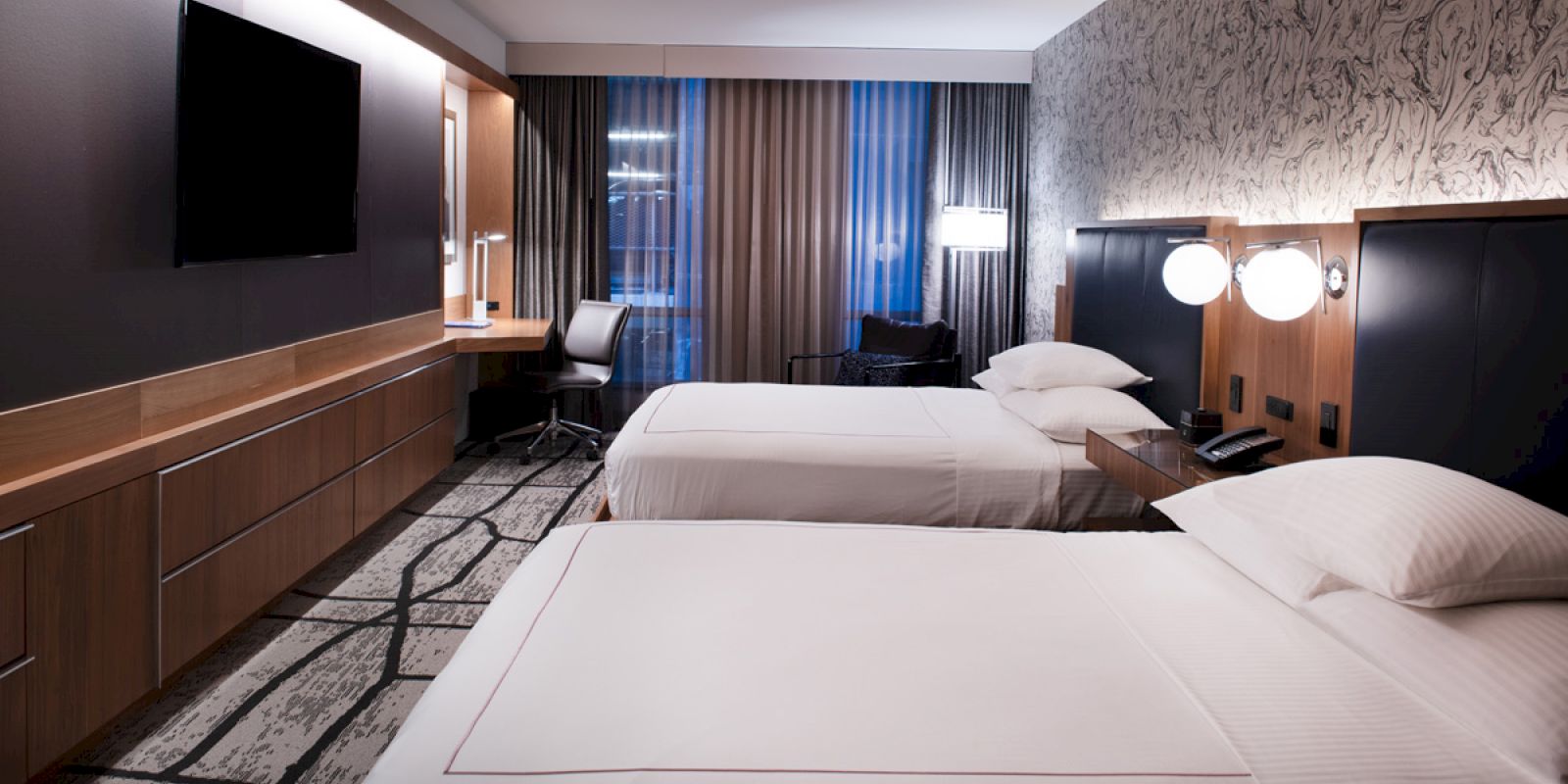 A modern hotel room with two neatly made beds, a flat-screen TV, a desk with a chair, and stylish lighting and decor ending the sentence.