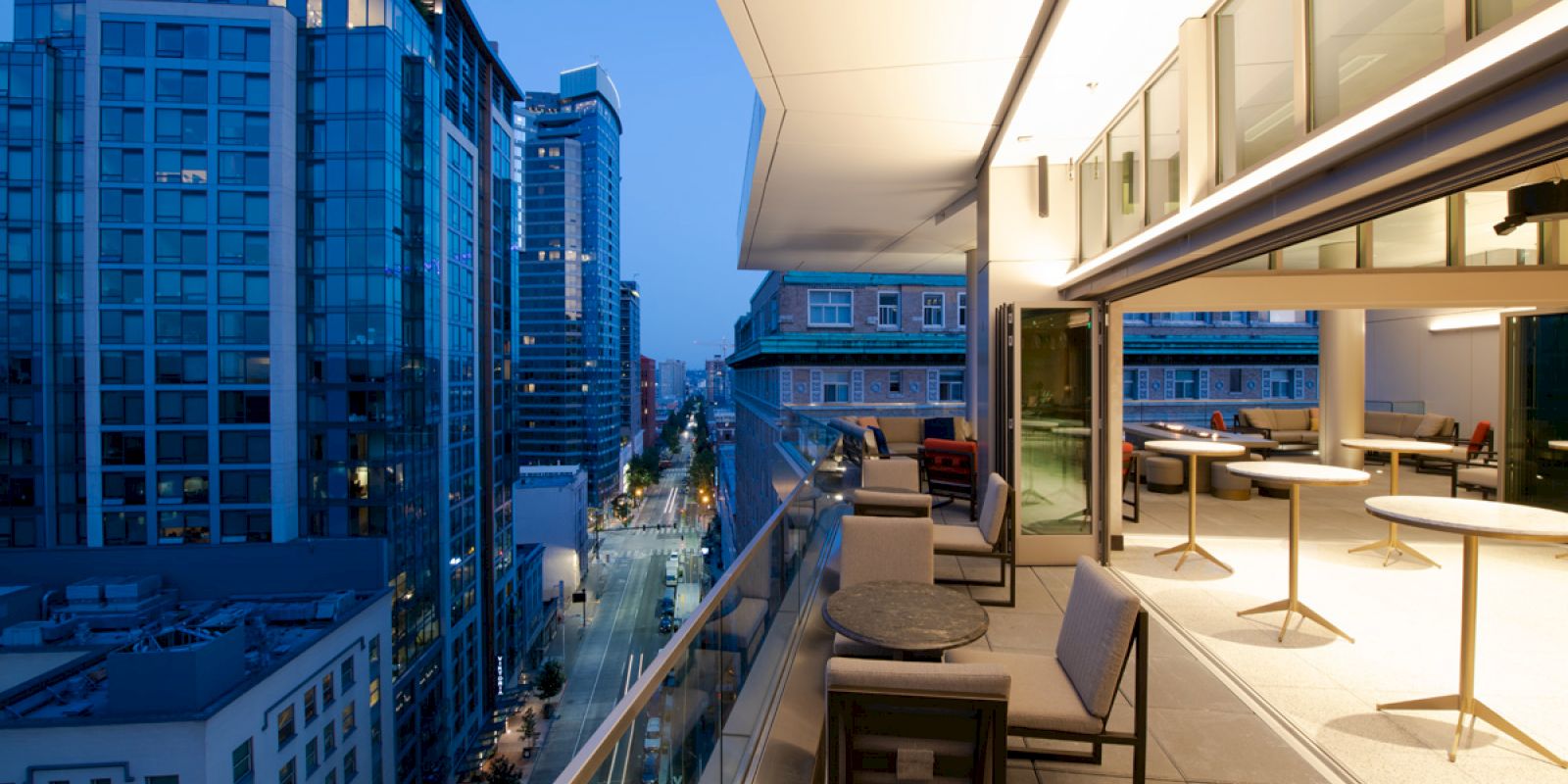 The image shows a modern cityscape with tall buildings and a large balcony with seating and tables, featuring a night-time view.