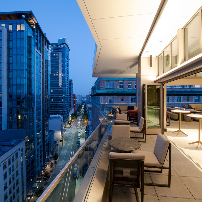 A modern balcony with tables and chairs overlooks a bustling city street lined with tall buildings at dusk.