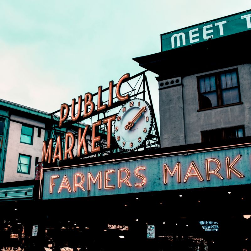 This image shows the entrance of a public farmers market with neon signs and a clock, located in an urban environment with nearby buildings.