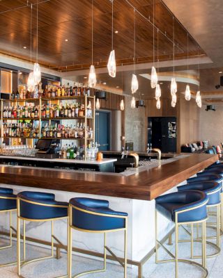 A modern bar with a wooden counter, blue stools, a wide selection of bottled drinks, overhead lighting, and a lounge area with sofas and chairs.