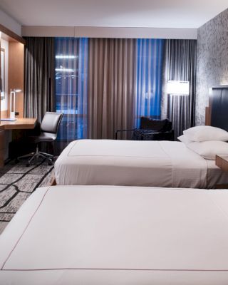 A modern hotel room with two neatly made beds, a wall-mounted TV, a desk with a chair, lamps, and patterned carpet flooring.