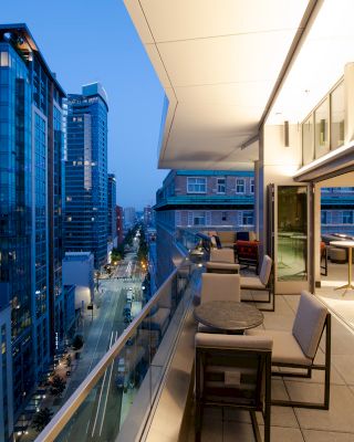 A modern balcony with seating overlooks a cityscape filled with tall buildings at dusk. The setting is elegant and lit with warm lights.