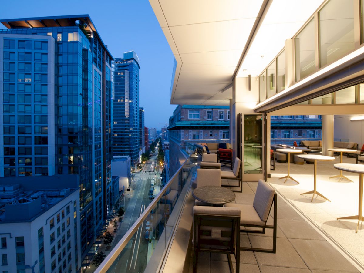 A modern cityscape at dusk featuring tall buildings, viewed from a balcony with seating and tables near a sleek, open space indoors.