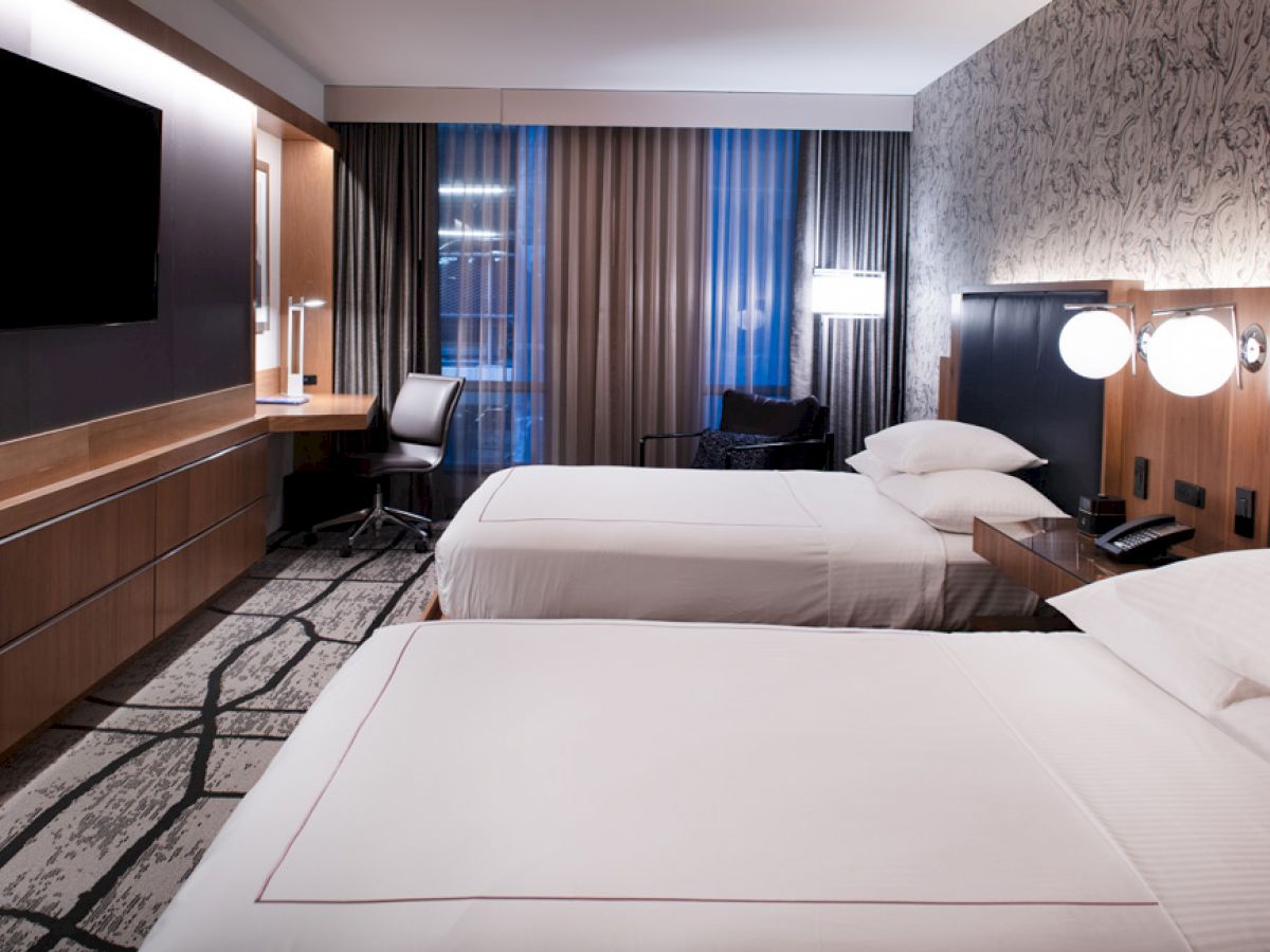A modern hotel room featuring two neatly made double beds, a large flat-screen TV, a desk with a chair, and stylish decor with patterned wallpaper.