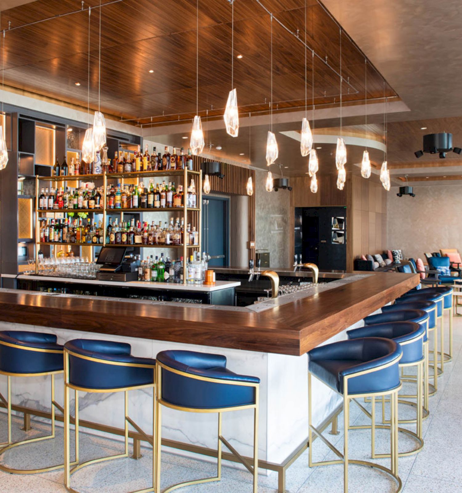 A modern bar with a wooden counter, blue bar stools, hanging lights, and a variety of bottles on shelves. The seating area is stylishly designed.