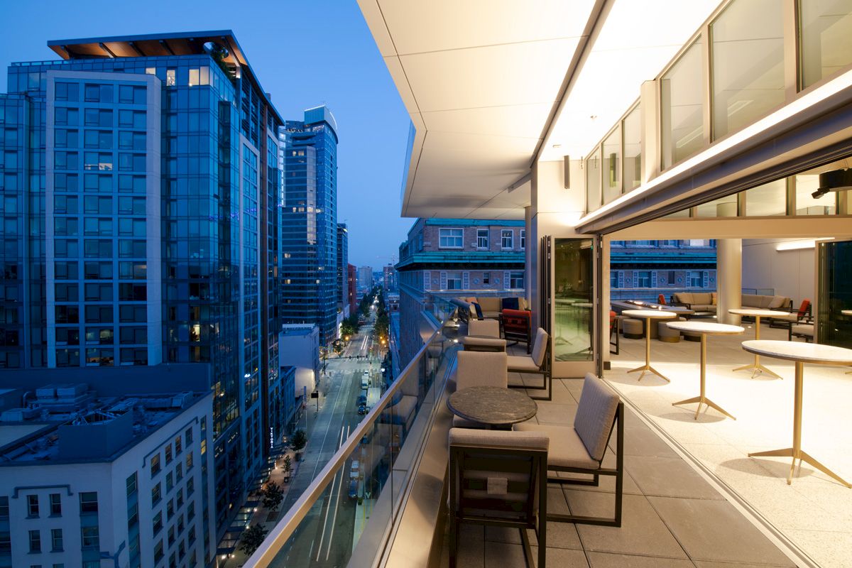 The image shows a modern balcony with seating and tables overlooking a city skyline at dusk, with high-rise buildings and a street below.