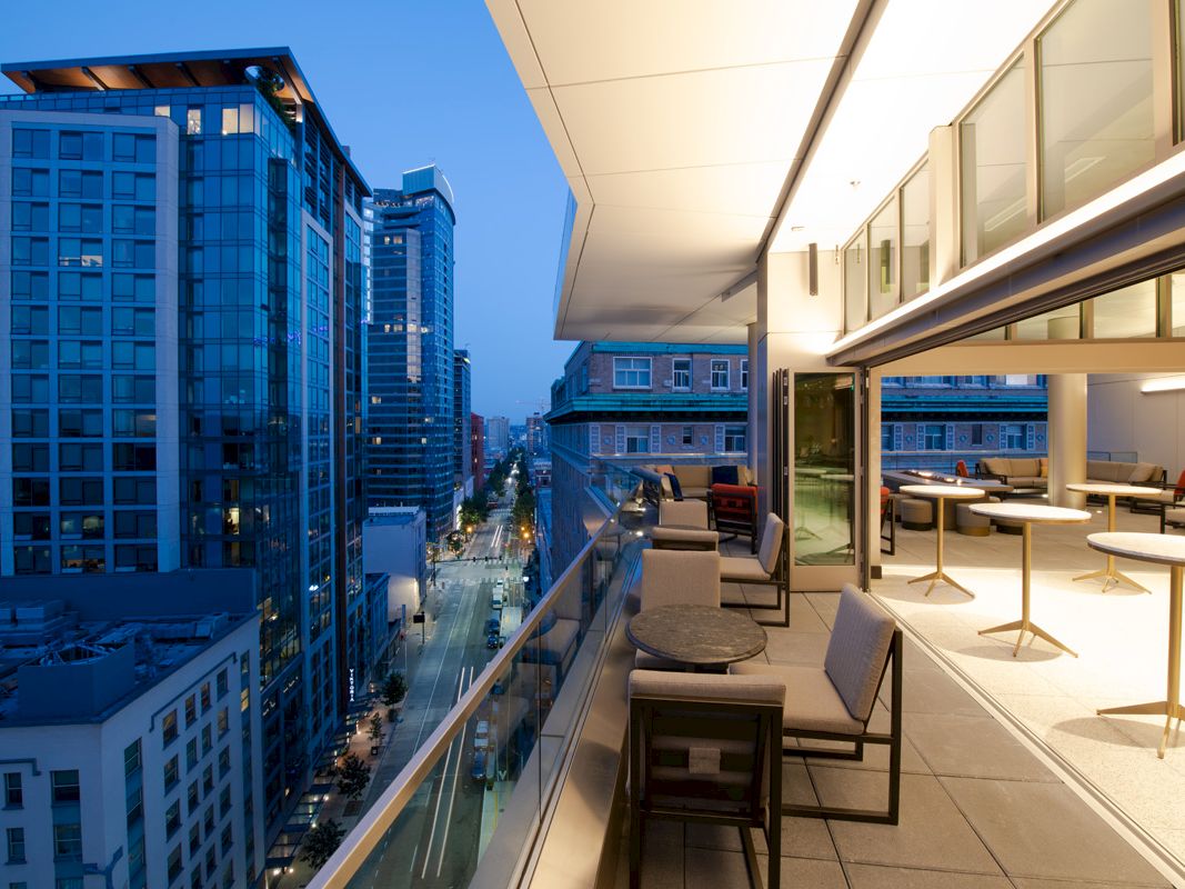 The image shows a modern balcony with seating and tables overlooking a city skyline at dusk, with high-rise buildings and a street below.