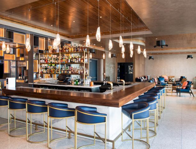 The image shows a modern bar with a wooden ceiling, contemporary lighting, a well-stocked bar, and blue bar stools arranged around a large counter.
