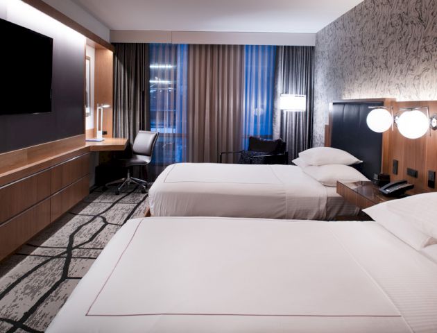 A modern hotel room with two beds, a wall-mounted TV, a desk, and a chair, with curtains and a patterned wallpaper.