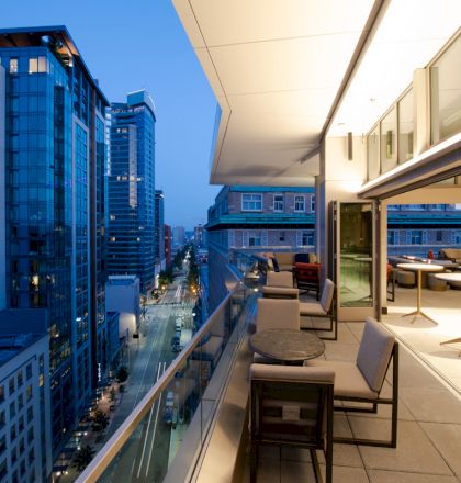 The image shows a modern urban balcony at dusk, featuring seating, tables, and city skyscrapers in the background, with street view below, ending the sentence.