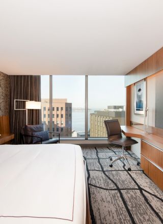 A modern hotel room features a large bed, wall art, a desk with a chair, and a large flat-screen TV, with a view of buildings outside the window.