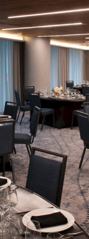 The image depicts a modern banquet hall set up for an event with round tables, chairs, elegant table settings, and floral centerpieces.