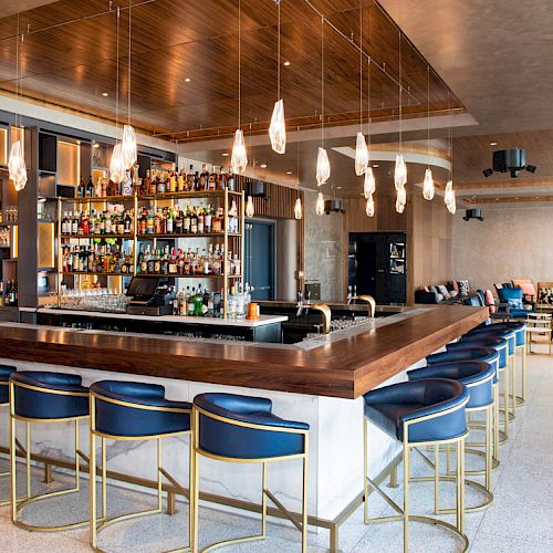 A modern bar with a wooden counter, blue bar stools, hanging lights, and a well-stocked shelf of liquor bottles behind the bar.