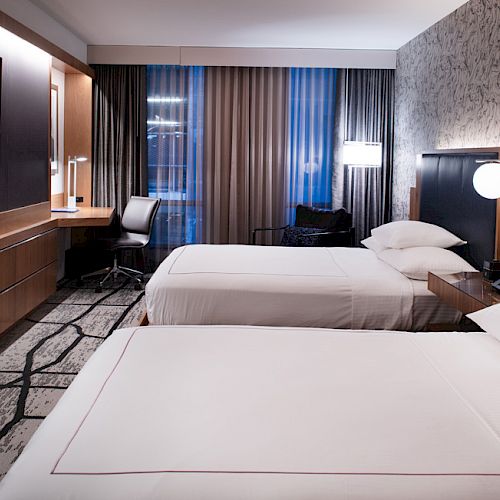 The image shows a modern hotel room with two beds, wall-mounted TV, desk with chair, and soft lighting from bedside lamps and windows.