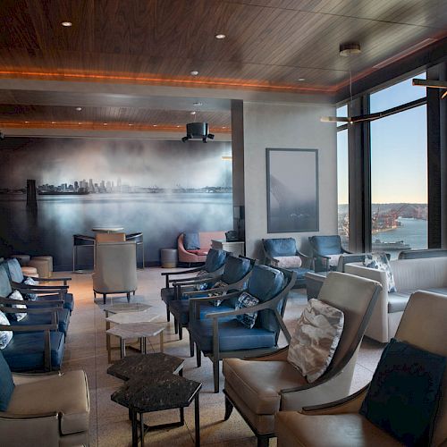 The image shows a modern lounge with several chairs, tables, large windows offering a scenic view of water and a Ferris wheel, and a cityscape mural.