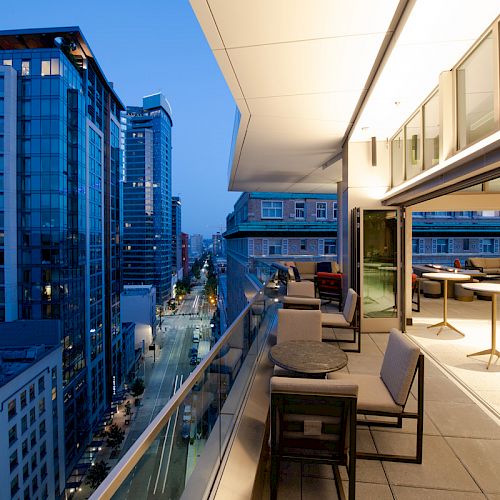 The image shows a modern balcony with furniture, overlooking a cityscape with tall buildings. It's evening, and the city lights are visible.
